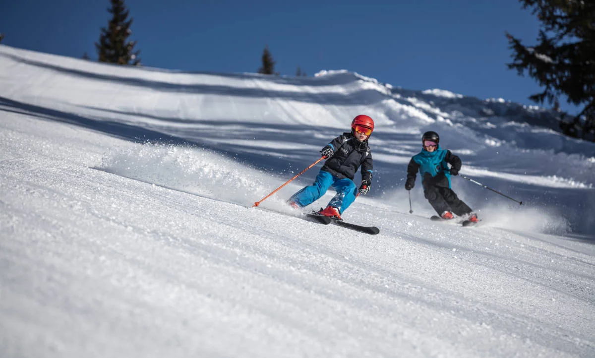Kinder beim Skifahren – Ski-Leasing & Skitauschsystem Frankfurt Kind fährt mit Skiern eine verschneite Piste hinunter, begleitet von einem Erwachsenen – Darstellung für Ski-Leasing für Kinder in Frankfurt