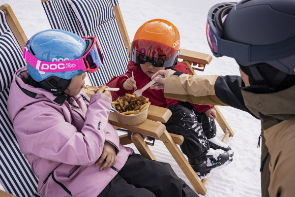 Drei Kinder mit Skihelmen entspannen im Schnee und teilen Snacks – ideal für Ski Leasing Kinder und Skitauschsysteme