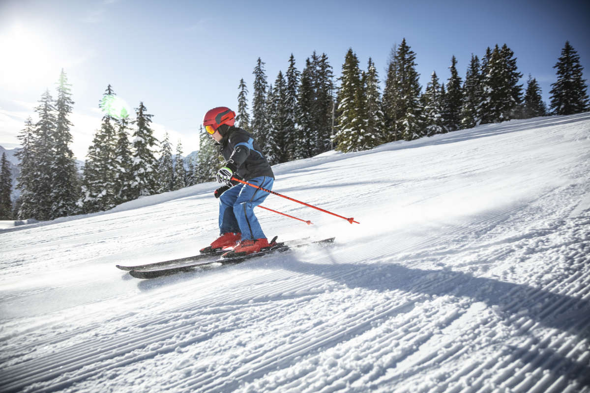 Kind fährt mit roten Skischuhen und Helm eine sonnige Skipiste hinunter – Bild für Ski-Leasing für Kinder in Frankfurt